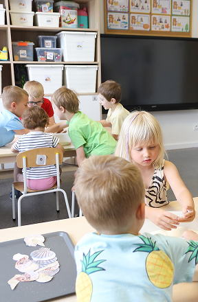 Kilkoro małych dzieci siedzi przy stolikach w sali lekcyjnej zaangażowanych w zajęcia plastyczne i rzemieślnicze, z pojemnikami do przechowywania i dużym ekranem w tle.