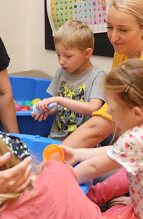 Chłopiec i dwie dziewczynki bawią się plastikowymi piłkami i pojemnikami w niebieskim pojemniku sensorycznym, podczas gdy dorosły nadzoruje ich pracę.