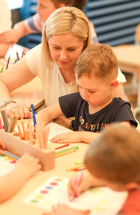 Kobieta pomaga małemu chłopcu w kolorowaniu przy stole, w otoczeniu innych dzieci i kolorowych kredek.