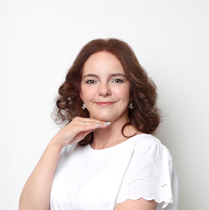 Kobieta z kręconymi brązowymi włosami w białej bluzce pozuje na białym tle, opierając podbródek na dłoni i uśmiechając się do kamery.