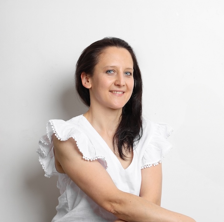 Kobieta z długimi ciemnymi włosami w białej bluzce z marszczonymi rękawami, siedząca na białym tle, lekko uśmiechnięta.