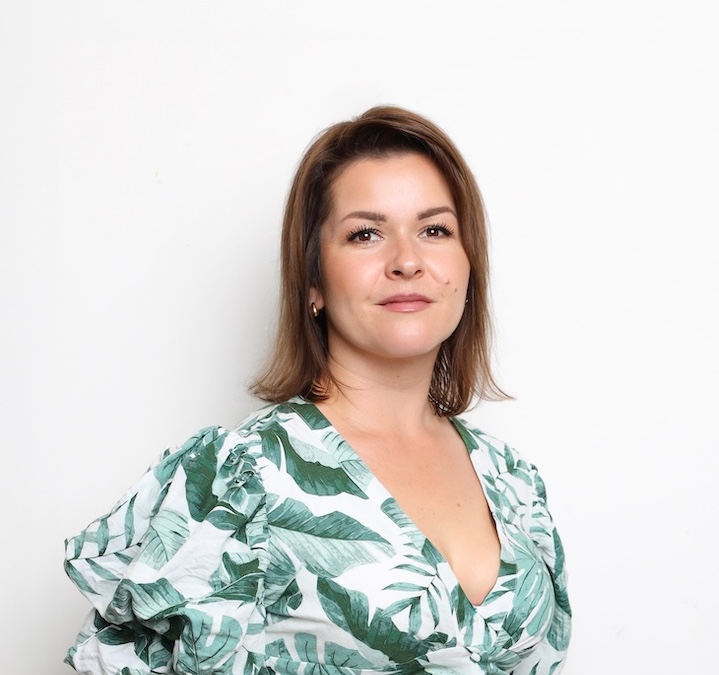 Kobieta z krótkimi brązowymi włosami ubrana w biało-zieloną bluzkę w liście stoi na białym tle i patrzy w kamerę.