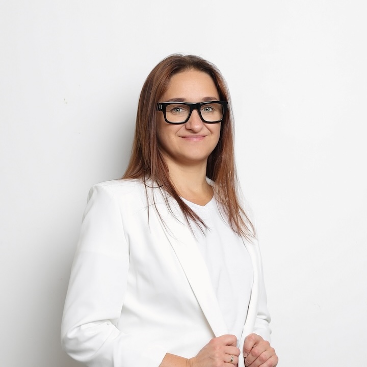 Kobieta z długimi brązowymi włosami w okularach i białej marynarce stoi na prostym białym tle, patrząc w kamerę i lekko się uśmiechając.
