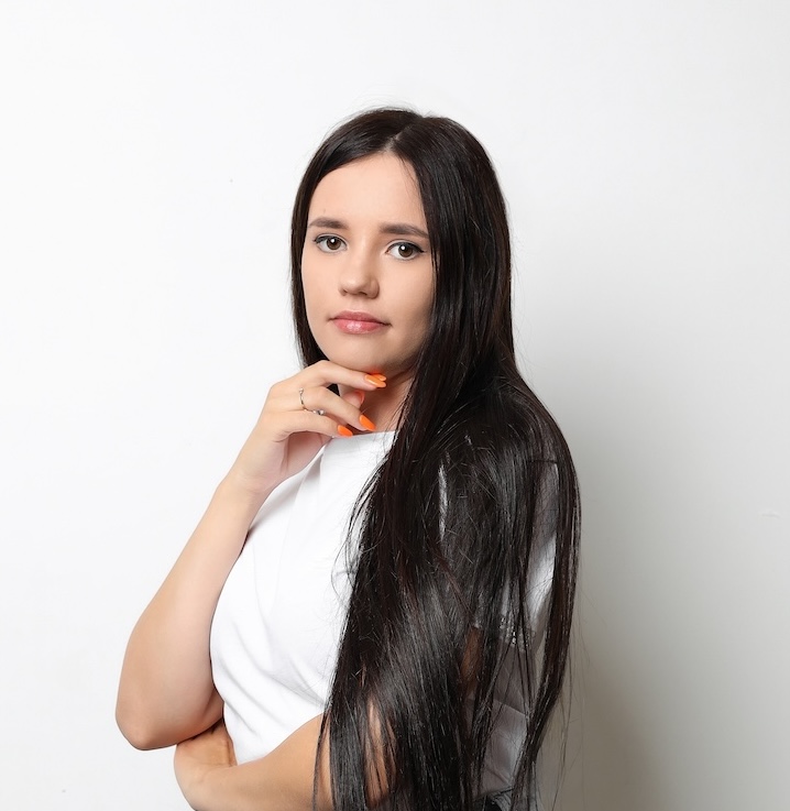Kobieta z długimi ciemnymi włosami, ubrana w białą koszulę, stoi na prostym białym tle, patrząc w kamerę z neutralnym wyrazem twarzy.
