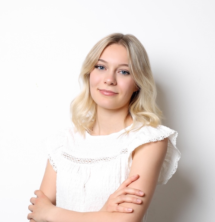 Młoda osoba z blond włosami i białą bluzką stoi na prostym białym tle, patrząc w kamerę z neutralnym wyrazem twarzy i skrzyżowanymi ramionami.