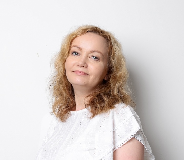 Kobieta z falującymi blond włosami i białą bluzką stoi na prostym białym tle, patrząc w kamerę z neutralnym wyrazem twarzy.