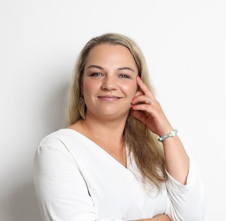 Kobieta z długimi blond włosami, ubrana w biały top, złote kolczyki i bransoletkę z koralików, pozuje na białym tle, uśmiechając się z ręką przy twarzy.