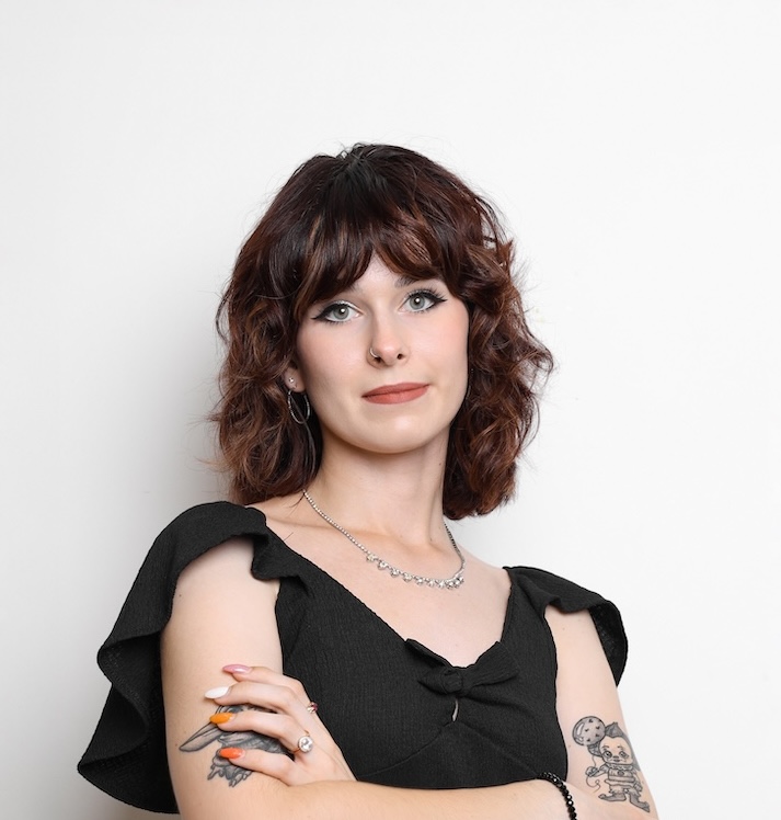 Osoba z falującymi brązowymi włosami i tatuażami na obu ramionach stoi na prostym białym tle, ubrana w czarną sukienkę i biżuterię, ze skrzyżowanymi rękami.