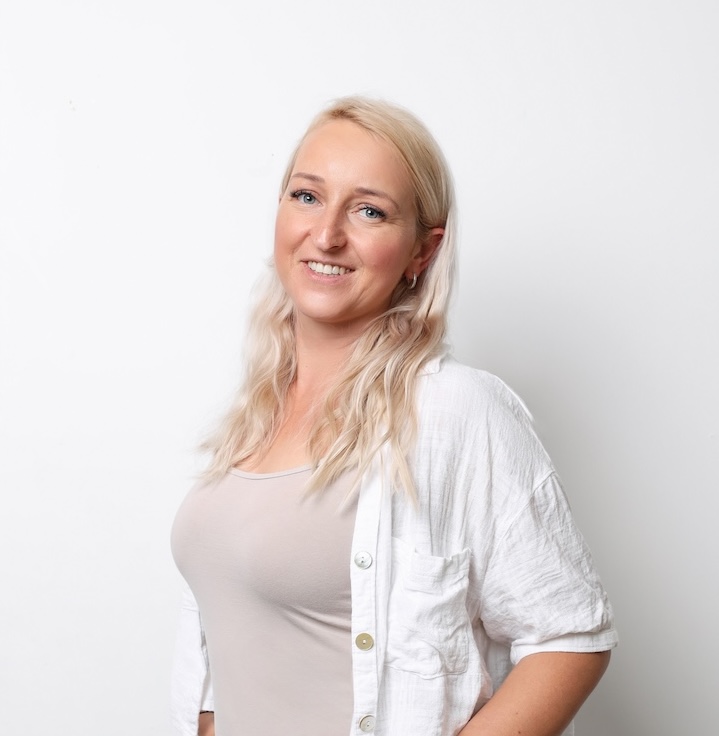 Kobieta z długimi blond włosami ubrana w jasnobeżowy top i białą koszulę zapinaną na guziki stoi przed białym tłem i uśmiecha się do kamery.