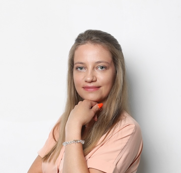 Kobieta z długimi blond włosami, ubrana w jasnoróżową koszulę i bransoletkę, pozuje z ręką przy brodzie na białym tle.