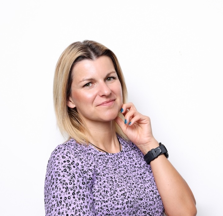 Kobieta z blond włosami sięgającymi ramion, ubrana w fioletową wzorzystą koszulę i czarny zegarek pozuje na białym tle.