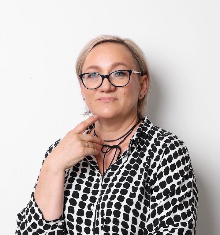 Kobieta z krótkimi blond włosami i okularami ubrana w czarno-białą wzorzystą koszulę pozuje na prostym białym tle, dotykając podbródka jedną ręką.