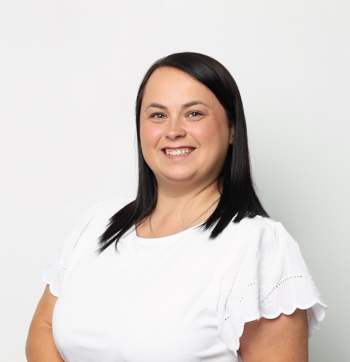 Kobieta z prostymi ciemnymi włosami, ubrana w białą bluzkę z krótkim rękawem, uśmiecha się do kamery na białym tle.