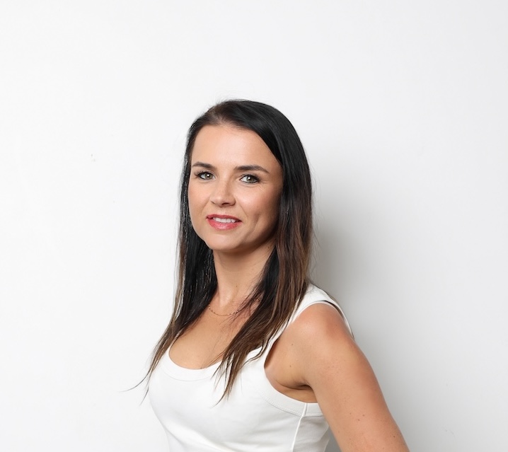 Kobieta z długimi ciemnymi włosami i białym topem bez rękawów pozuje na prostym białym tle, patrząc w kamerę z lekkim uśmiechem.
