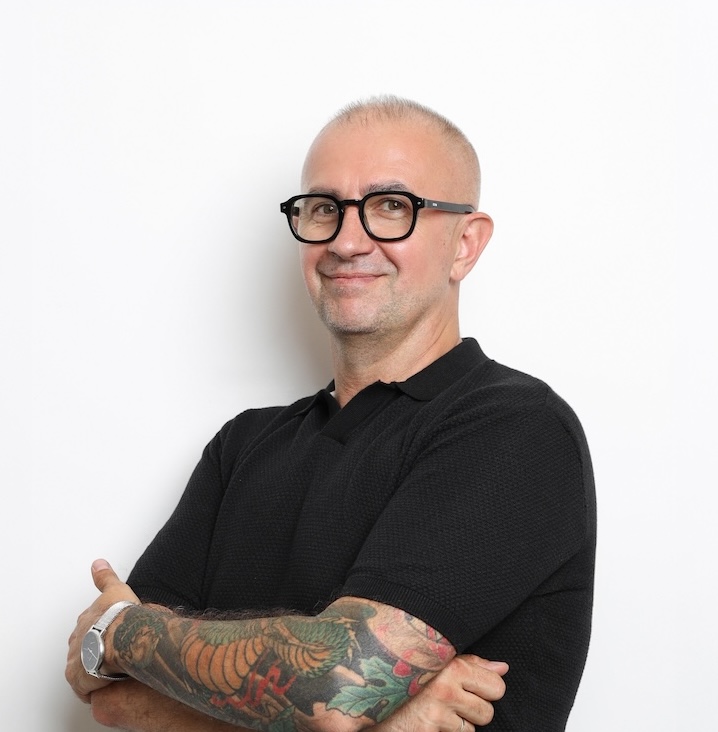 Mężczyzna w okularach, z krótkimi włosami i tatuażami na przedramionach stoi na białym tle, ubrany w czarną koszulkę polo z krótkim rękawem i uśmiecha się ze skrzyżowanymi ramionami.