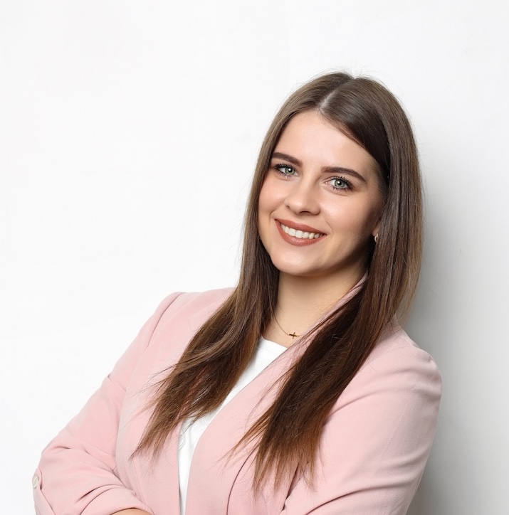 Kobieta z długimi brązowymi włosami w różowej marynarce i białej koszuli stoi na białym tle, uśmiechając się ze skrzyżowanymi ramionami.