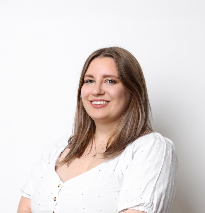 Kobieta z długimi brązowymi włosami, ubrana w białą bluzkę, uśmiecha się do kamery na białym tle.