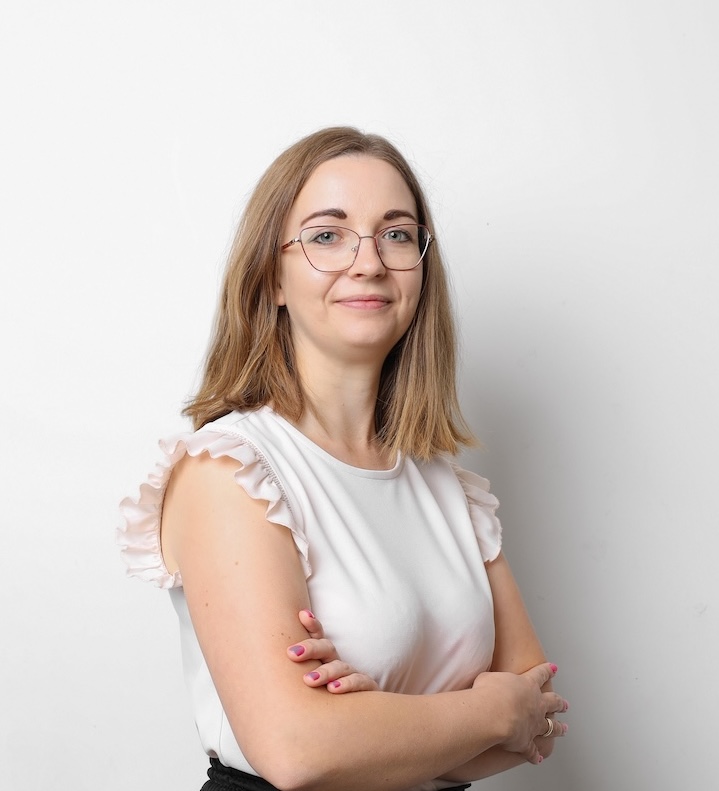 Kobieta w okularach i jasnobrązowych włosach do ramion, ubrana w białą bluzkę bez rękawów, stoi ze skrzyżowanymi ramionami na białym tle.