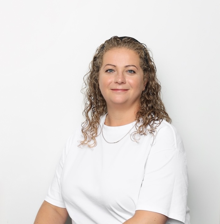 Kobieta z kręconymi włosami ubrana w białą koszulkę i cienki naszyjnik siedzi na prostym białym tle, twarzą do kamery i lekko się uśmiecha.