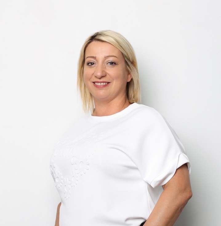 Kobieta o prostych blond włosach, ubrana w białą bluzkę z krótkim rękawem, stoi na białym tle i uśmiecha się do kamery.