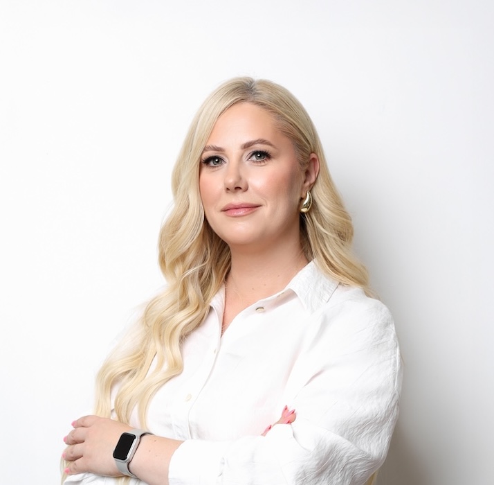 Kobieta z długimi blond włosami, ubrana w białą koszulę i elegancki zegarek, stoi na prostym białym tle ze skrzyżowanymi rękami.