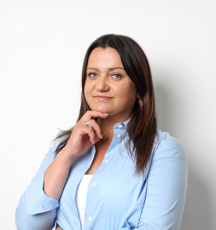 Kobieta z prostymi brązowymi włosami ubrana w jasnoniebieską koszulę zapinaną na guziki pozuje na prostym białym tle, opierając dłoń w zamyśleniu na brodzie.