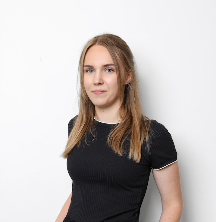 Młoda kobieta z długimi blond włosami ubrana w czarny top z krótkim rękawem stoi na prostym białym tle, patrząc w kamerę z neutralnym wyrazem twarzy.