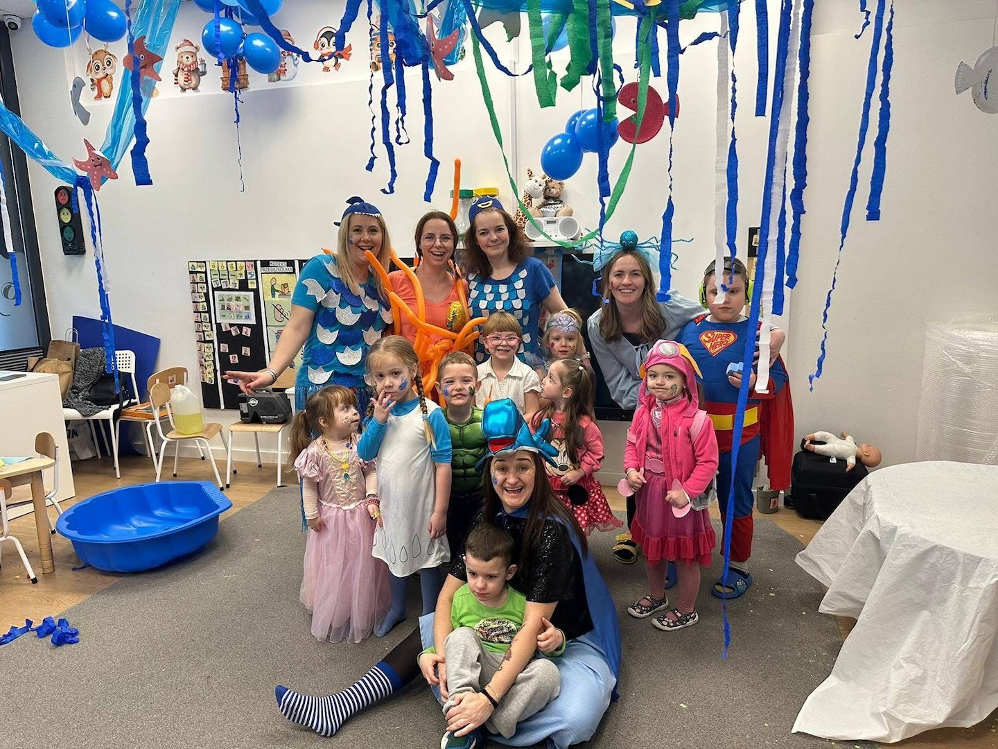 Grupa dzieci i dorosłych w kolorowych kostiumach pozuje razem w udekorowanym pokoju z serpentynami, balonami i różnymi tematycznymi grafikami ściennymi.