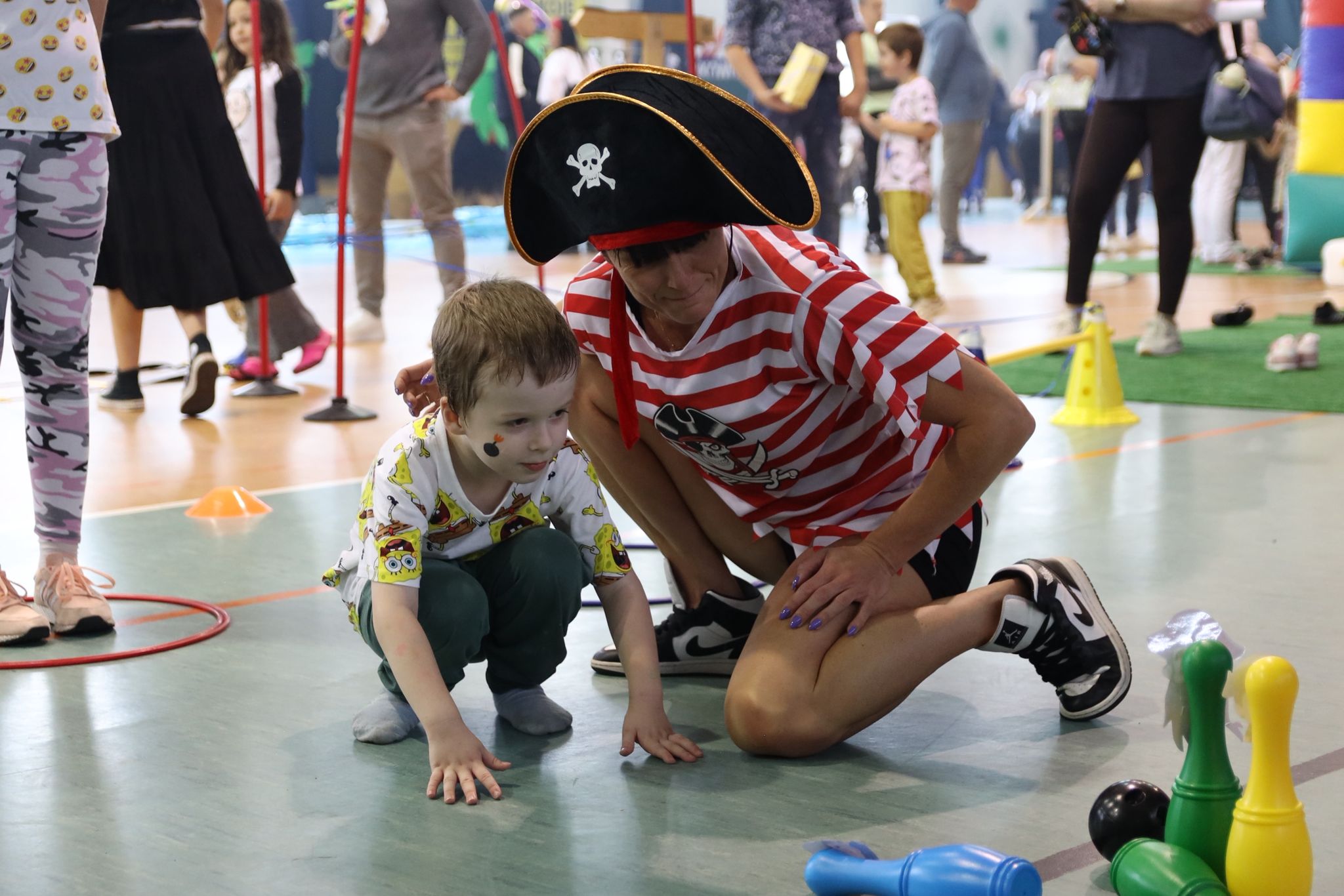 Dziecko i dorosły w stroju pirata kucają na podłodze w sali gimnastycznej, przygotowując się do gry plastikowymi kręgielniami; w tle widoczne są inne osoby i czynności.